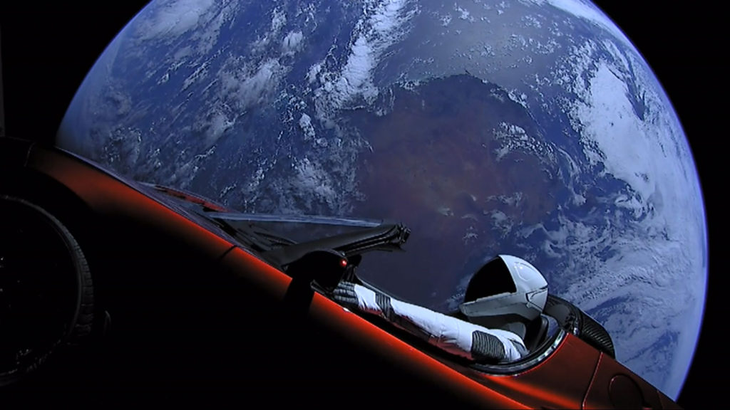 Veículo Tesla Roadster tripulado pelo boneco Starman no espaço, com o planeta Terra no fundo. O carro iniciou sua viagem pelas estrelas em fevereiro de 2018 no primeiro voo do foguete Falcon Heavy.